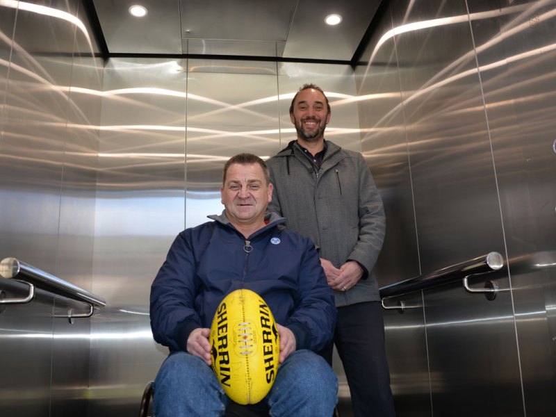 One wheelchair user with an AFL football and another man in a lift at North Hobart Football Club.