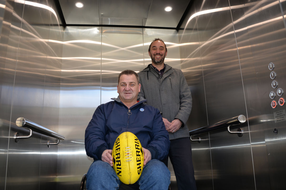 One wheelchair user with an AFL football and another man in a lift at North Hobart Football Club.